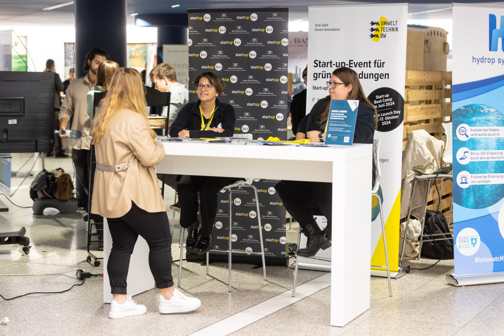 Im Bild ist ein Ausstellungstisch der Landesagentur Start-up BW zu sehen, an dem sich drei Personen unterhalten. Im Hintergrund ein Rollbanner, auf dem steht: Start-up Event für grüne Gründungen.